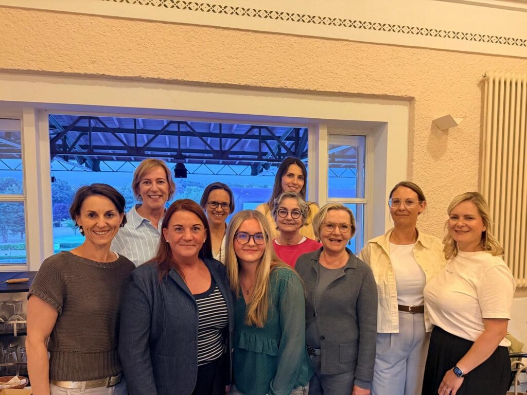 Zehn Frauen aus dem LLiT-Netzwerk stehen lächelnd zusammen in einem hellen Raum vor einem großen Fenster; draußen sind eine Stahlkonstruktion und Bäume im Abendlicht zu sehen.