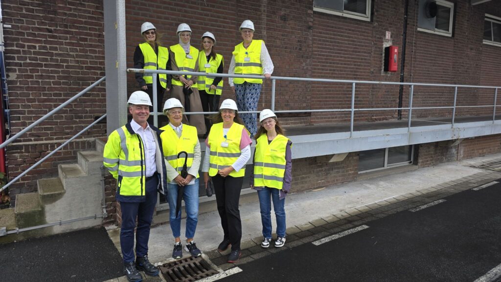 Gruppenfoto auf dem Werksgelände von Solenis in Krefeld: acht Frauen und ein Mann in gelben Warnwesten und weißen Schutzhelmen vor einem Backsteingebäude, vier davon auf einer Treppe mit Geländer.